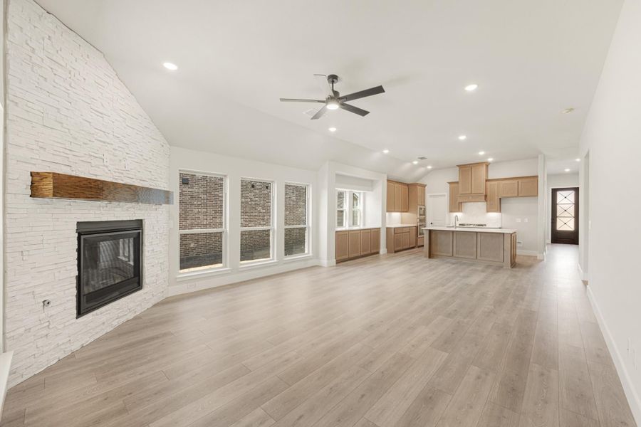 Open living area with white stone fireplace, wood mantel, and light hardwood floors open to kitchen with tan cabinets Open living area with white stone fireplace, wood mantel, and light hardwood floors open to kitchen with tan cabinets