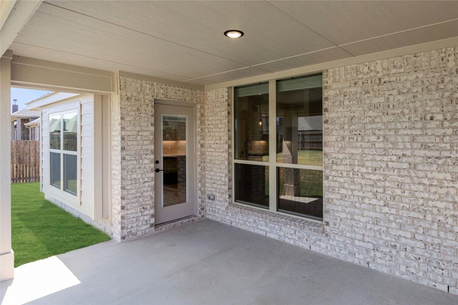 Covered back patio with concrete floor, white brick exterior, glass door, and large window overlooking green backyard