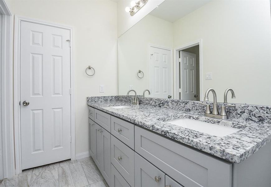 A clean, modern bathroom with double sinks, granite countertop, large mirror, and grey cabinets. White doors and tiled floor are visible.