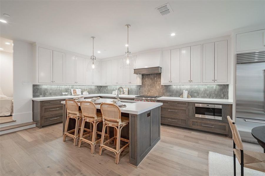 Kitchen featuring premium range hood, decorative light fixtures, an island with sink, stainless steel appliances, and white cabinets