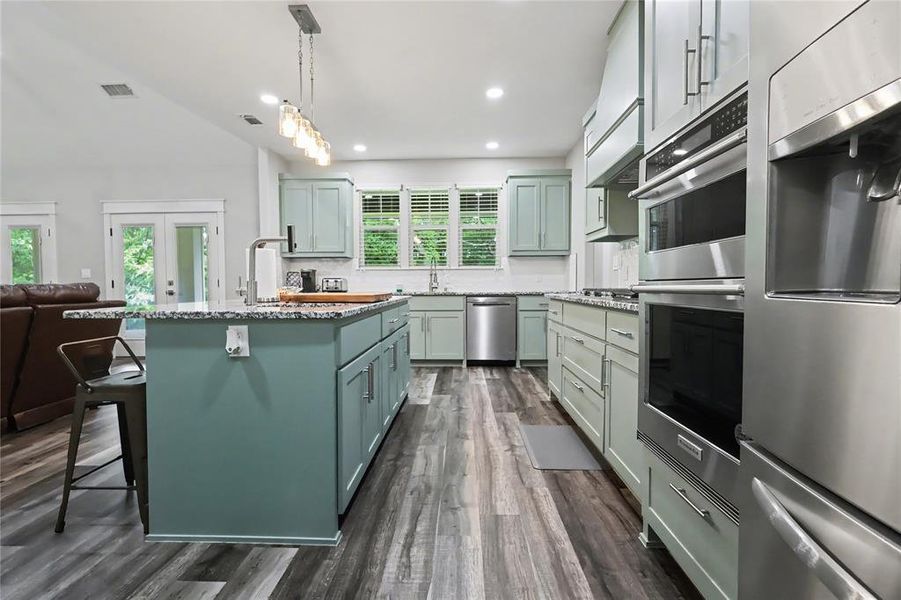 Spacious kitchen featuring a central island with a granite countertop, wood-finish flooring, stainless steel appliances, shaker-style cabinetry, and recessed lighting