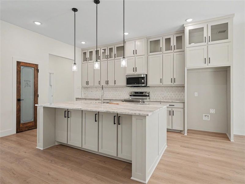 Kitchen featuring an expansive island with a stone countertop, abundant white cabinetry including glass-front uppers, a patterned tile backsplash, and wood-finish flooring Kitchen featuring an expansive island with a stone countertop, abundant white cabinetry including glass-front uppers, a patterned tile backsplash, and wood-finish flooring
