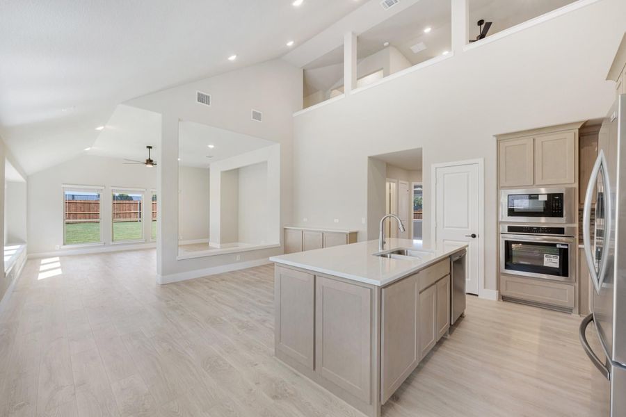 Open-concept kitchen with center island, stainless steel appliances, and high ceilings overlooking living area with light wood floors
