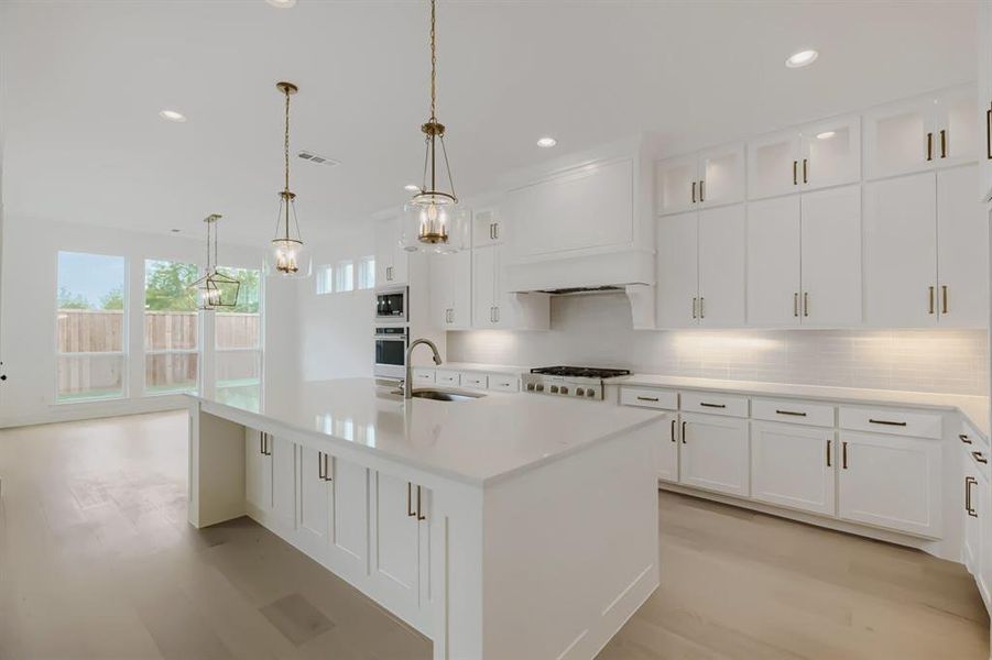 Spacious kitchen featuring an expansive island with a sink, white cabinetry with gold-toned hardware, stainless steel appliances including a built-in oven and gas range, and wood-finish flooring Spacious kitchen featuring an expansive island with a sink, white cabinetry with gold-toned hardware, stainless steel appliances including a built-in oven and gas range, and wood-finish flooring