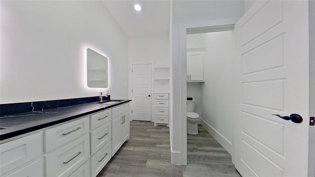 Spacious bathroom featuring a dual vanity with white cabinetry and dark countertops, an illuminated mirror, and wood-finish flooring Spacious bathroom featuring a dual vanity with white cabinetry and dark countertops, an illuminated mirror, and wood-finish flooring