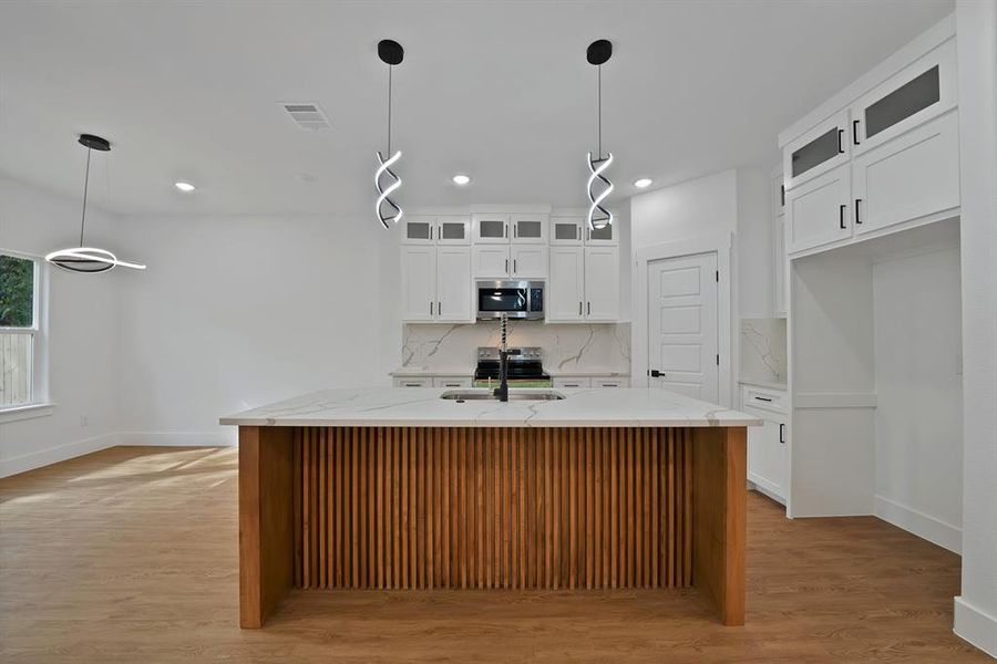 Kitchen featuring decorative light fixtures, light stone counters, glass insert cabinets, a center island with sink, and tasteful backsplash