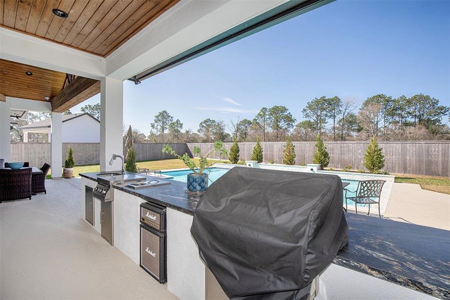 This photo showcases a spacious outdoor patio with a built-in grill and kitchen area, overlooking a pool. The area is surrounded by a privacy fence and landscaped with trees, offering a serene backyard setting.