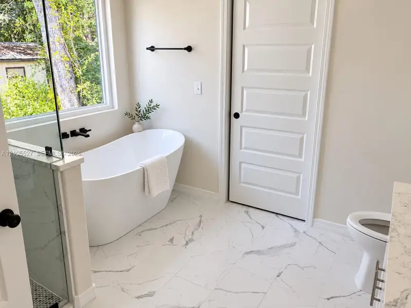 Freestanding soaking tub with matte black fixtures, framed window, and marble-look floor tiling Freestanding soaking tub with matte black fixtures, framed window, and marble-look floor tiling