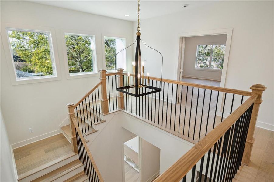 The upstairs landing exudes elegance and natural light, creating a bright and airy transition space. Oversized windows frame picturesque views, while the modern chandelier adds a bold yet refined touch. Wood accents on the stair railing and posts beautifully complement the iron balusters, blending warmth with contemporary design. The open layout connects seamlessly to surrounding rooms, highlighting the thoughtful flow and functionality.