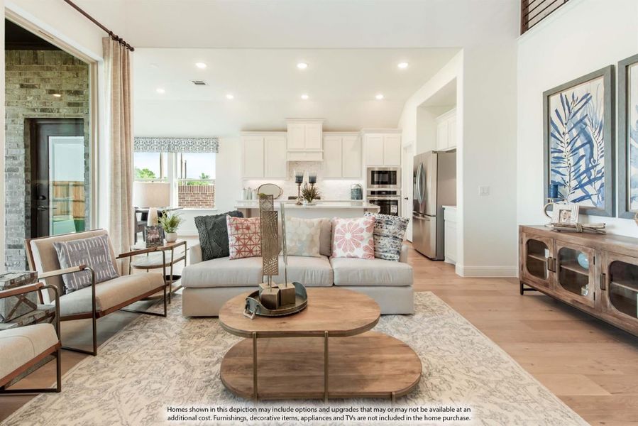Open-concept living room with light wood floors flowing into a white kitchen with stainless steel appliances and large windows.