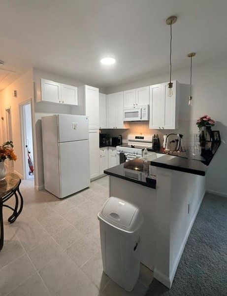 This photo showcases a compact, well-lit kitchen with white cabinetry and appliances, including a dishwasher and microwave. It features a small peninsula with pendant lighting, offering a cozy space for meal prep and dining. The kitchen opens into a hallway, enhancing accessibility and flow.