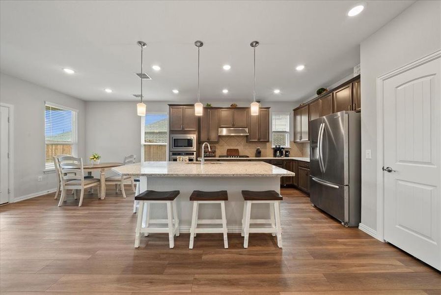 Kitchen with luxury vinyl plank floors, a center island with sink, appliances with stainless steel finishes, decorative light fixtures, and light stone counters.