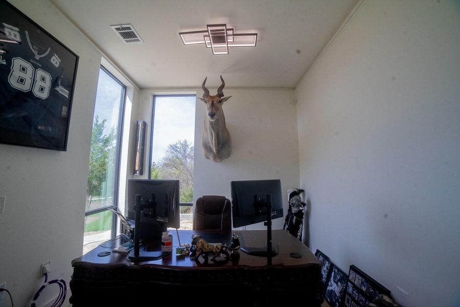Boost your productivity in this expansive home office, where massive windows frame tranquil greenery views and bathe the room in invigorating natural light. A statement unique ceiling light illuminates the sleek wooden desk, while high-end architectural details and ample space for a dual-monitor setup create a sophisticated, modern environment tailored for the professional