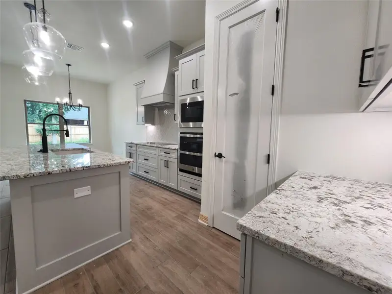 Kitchen featuring a large island with an integrated sink and matte black faucet, light-toned cabinetry, stainless steel built-in appliances, and light wood-finish flooring