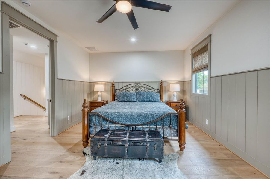 Bedroom featuring wood-finish flooring, half-wall wainscoting, a ceiling fan with integrated lighting, and a window with woven shades