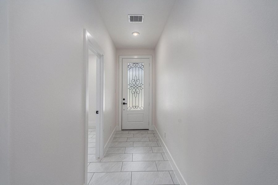 This photo shows a bright, modern hallway with light tile flooring and neutral walls. A decorative glass front door adds elegance, while a side opening leads to another room.