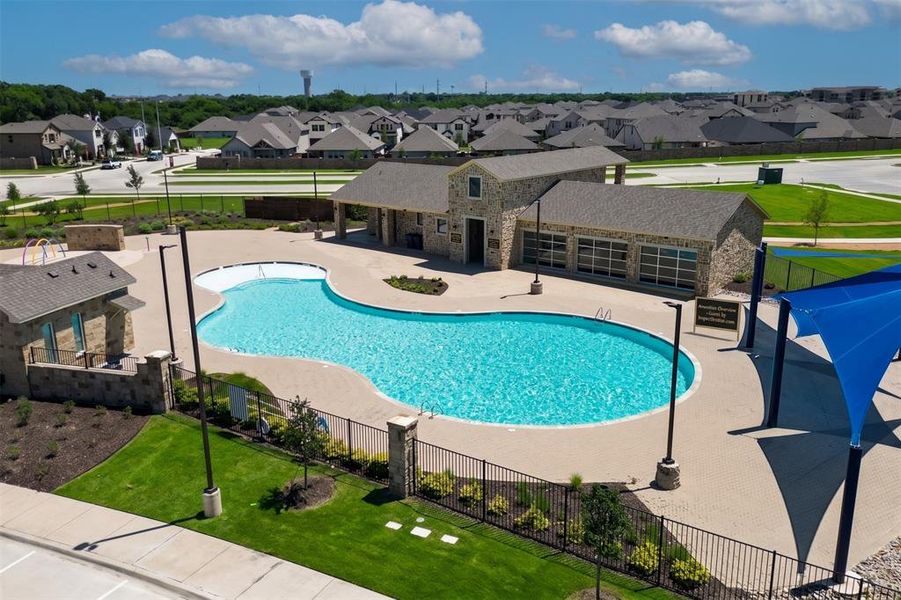 Community pool with integrated splash pad, featuring a stone-clad amenity building, paver decking, and dedicated shade structures