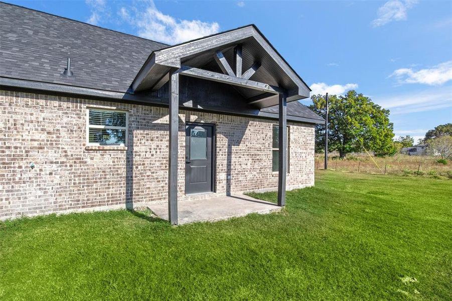 Property entrance with a shingled roof, a patio, a lawn, and brick siding