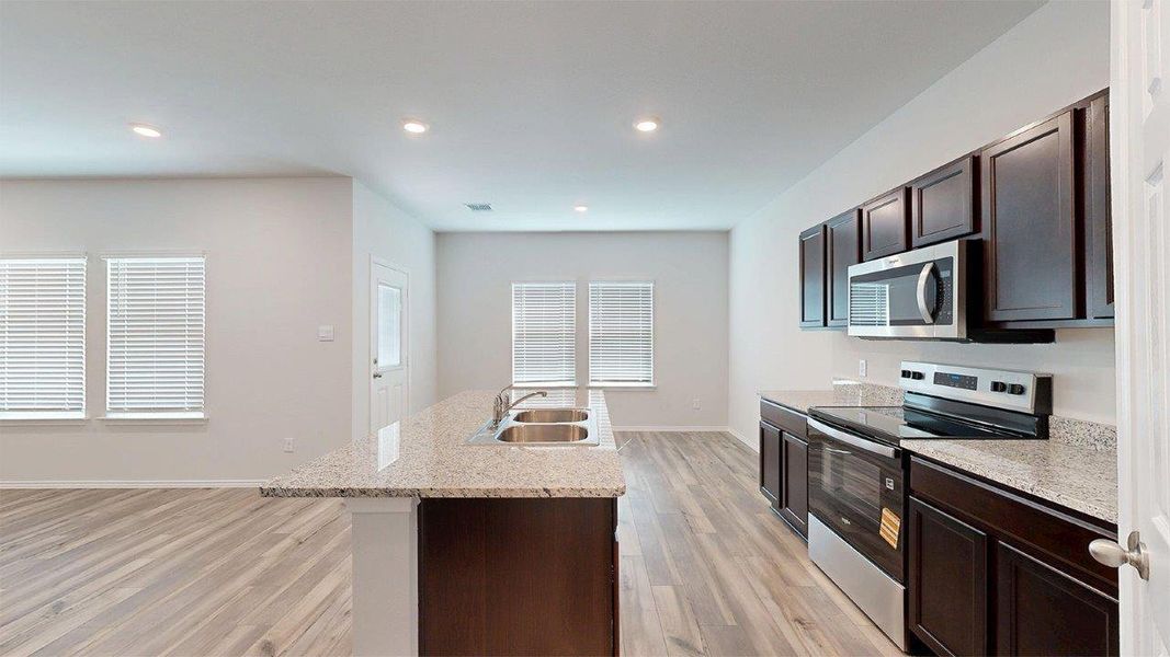 Open-concept kitchen featuring dark wood cabinetry, light granite countertops, stainless steel appliances, a double basin sink, and wood-finish flooring Open-concept kitchen featuring dark wood cabinetry, light granite countertops, stainless steel appliances, a double basin sink, and wood-finish flooring