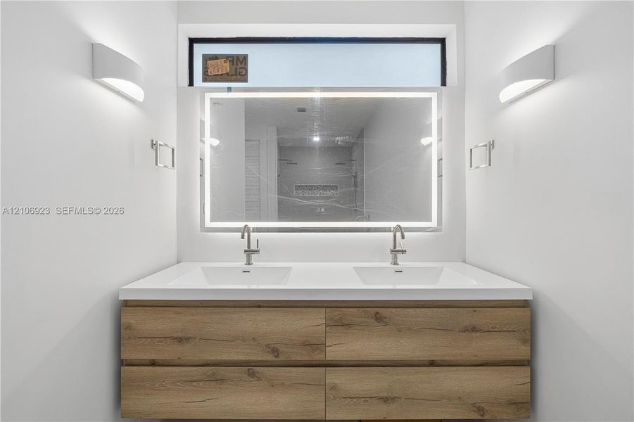 Double vanity with wood-finish cabinetry, solid surface countertop, integrated dual sinks, LED-backlit mirror, and wall-mounted sconces Double vanity with wood-finish cabinetry, solid surface countertop, integrated dual sinks, LED-backlit mirror, and wall-mounted sconces