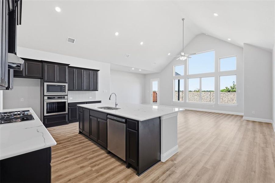 Kitchen featuring a high ceiling, an island with sink, open floor plan, light wood-style flooring, and light stone counters Kitchen featuring a high ceiling, an island with sink, open floor plan, light wood-style flooring, and light stone counters