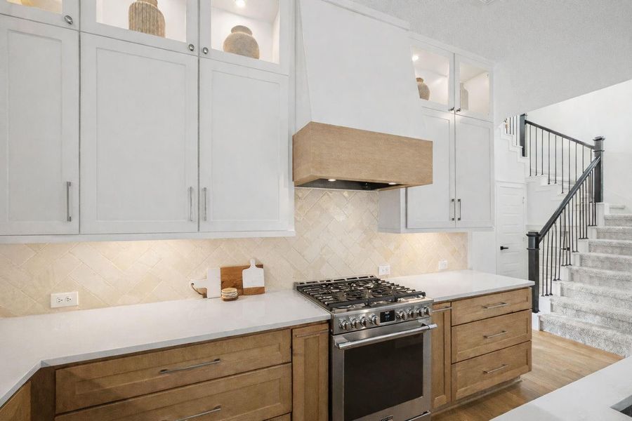 Statement cooking space anchored by a professional-style gas range, custom vent hood, and expansive prep surfaces. Tall cabinetry with lit display niches draws the eye upward, while the warm wood and soft tile backsplash add texture and character to this beautifully designed kitchen focal point.