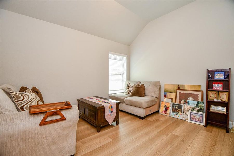 Cozy, well-lit room with light wood flooring, neutral walls, and a vaulted ceiling. Features comfortable seating, a small wooden coffee table, and a corner display of artwork and books. Ideal for a reading nook or relaxation space.