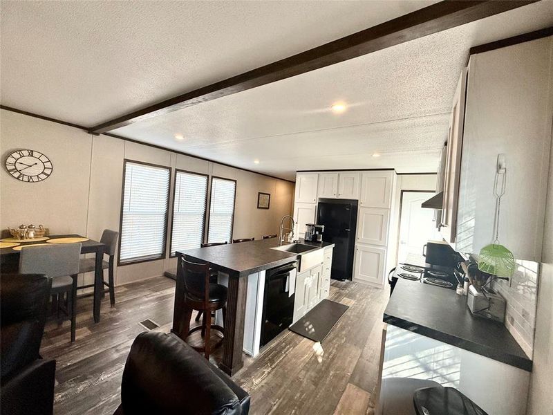 Kitchen with an island with sink, a textured ceiling, beamed ceiling, white cabinetry, and dark countertops