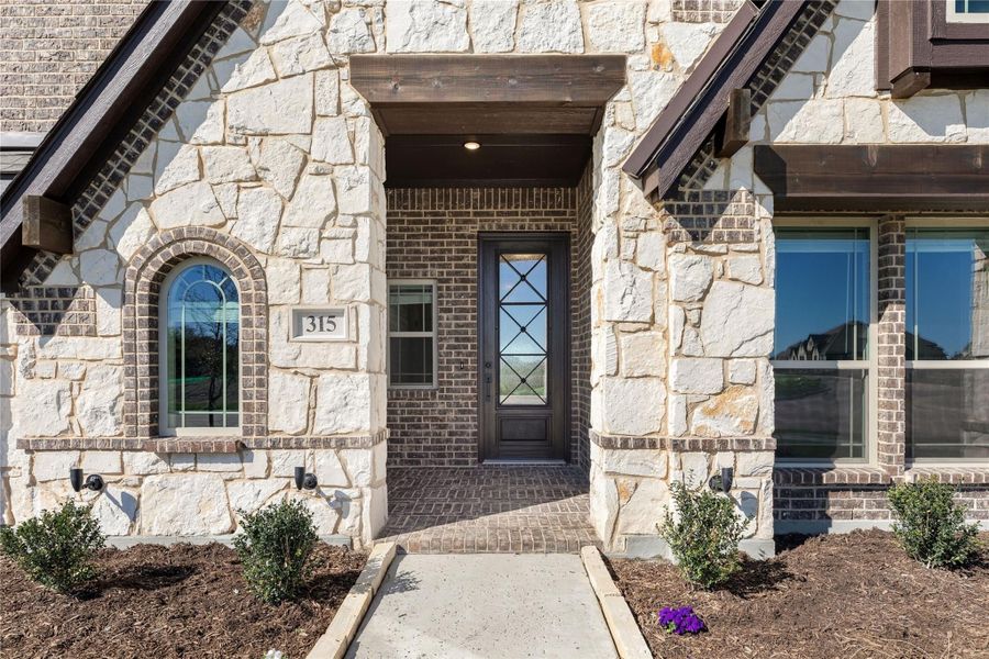 Home entrance with stone and brick exterior, dark front door with glass panels, and brick walkway Home entrance with stone and brick exterior, dark front door with glass panels, and brick walkway