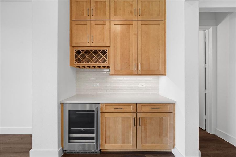 Built-in beverage station featuring light wood cabinetry, a dedicated wine rack, white subway tile backsplash, a light-toned countertop, and a stainless steel under-counter beverage refrigerator