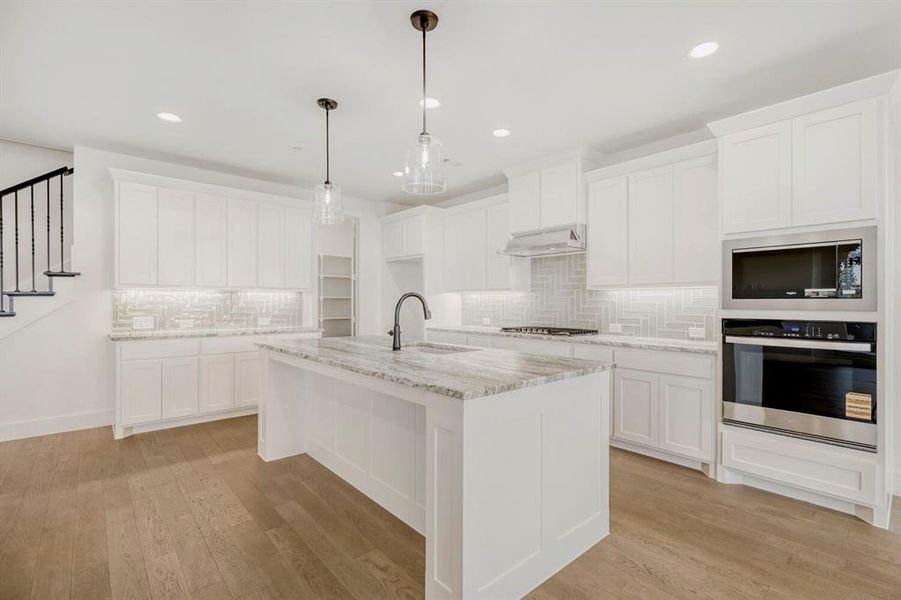 Open-concept kitchen featuring an oversized island with a built-in sink, stainless steel appliances, herringbone tile backsplash, recessed lighting, and wood-finish flooring