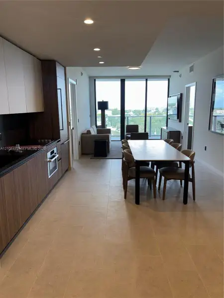 Modern kitchen featuring wood-finish cabinetry, built-in stainless steel oven, stone-surface countertops, recessed ceiling lighting, and floor-to-ceiling glass doors opening to a private balcony Modern kitchen featuring wood-finish cabinetry, built-in stainless steel oven, stone-surface countertops, recessed ceiling lighting, and floor-to-ceiling glass doors opening to a private balcony