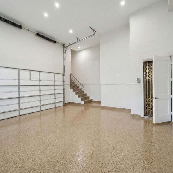 An oversized garage featuring durable epoxy flooring, high ceilings, and a modern paneled garage door system. A private elevator provides direct access to the home, while an interior staircase offers an additional entry point. Bright recessed lighting and clean finished walls create a polished, functional space designed for both convenience and storage.