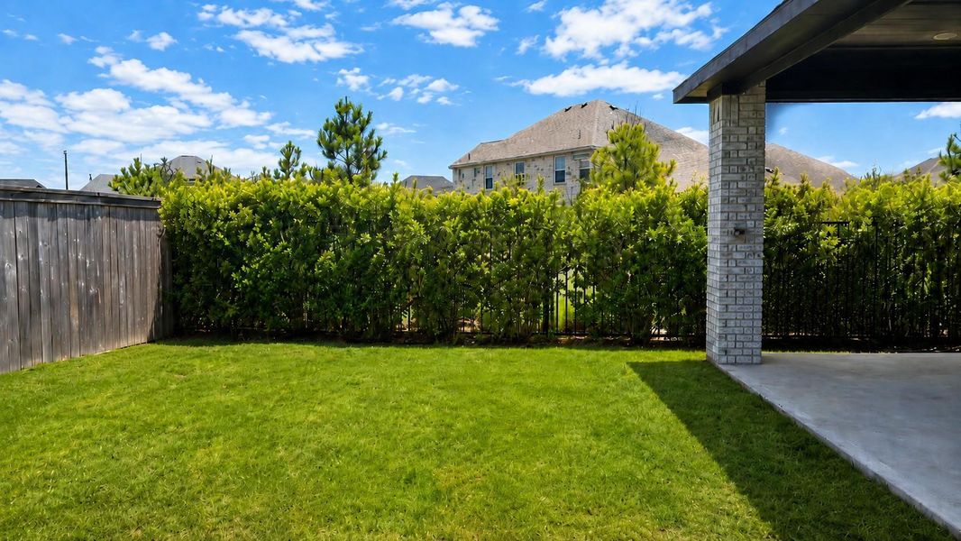 A side view of the backyard shows a green lawn bordered by a thick privacy hedge line. A wooden privacy fence runs along the perimeter next to the covered patio area.