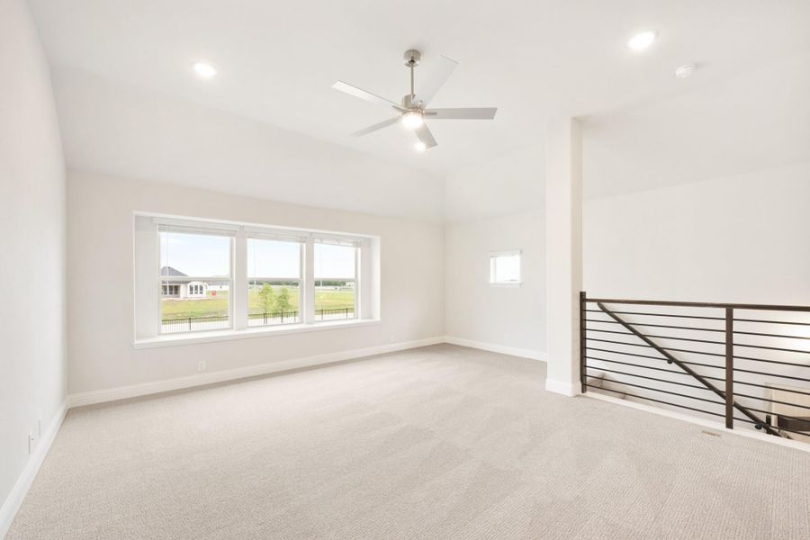 Open upstairs loft with carpet, ceiling fan, large window, and metal stair railing overlooking lower level