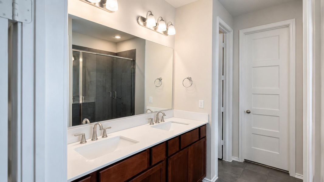 The bathroom includes a double vanity with a white countertop, two undermount sinks, and dark wood lower cabinets. A large wall mirror reflects a walk-in tile shower with glass doors.