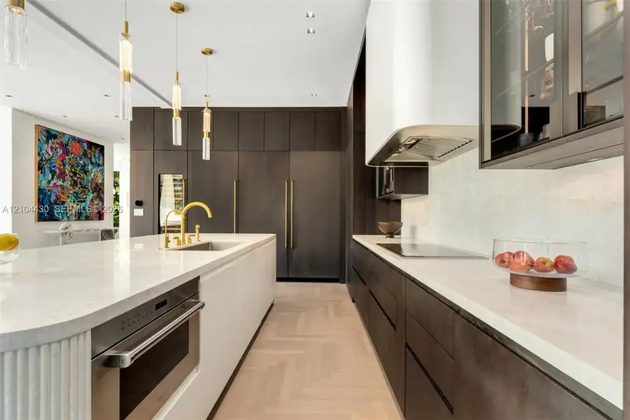 Gourmet kitchen featuring custom dark cabinetry, quartz countertops, a central island with a fluted pillar detail, herring-bone pattern wood-finish flooring, and integrated stainless steel appliances