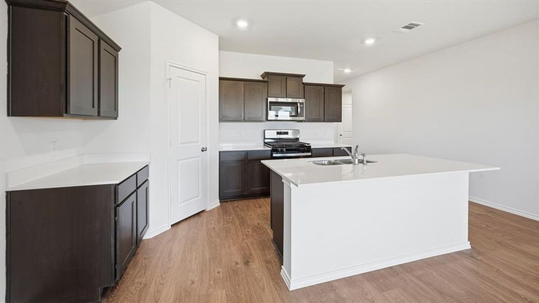 Kitchen featuring appliances with stainless steel finishes, a center island with sink, dark brown cabinetry, light stone countertops, and light wood finished floors