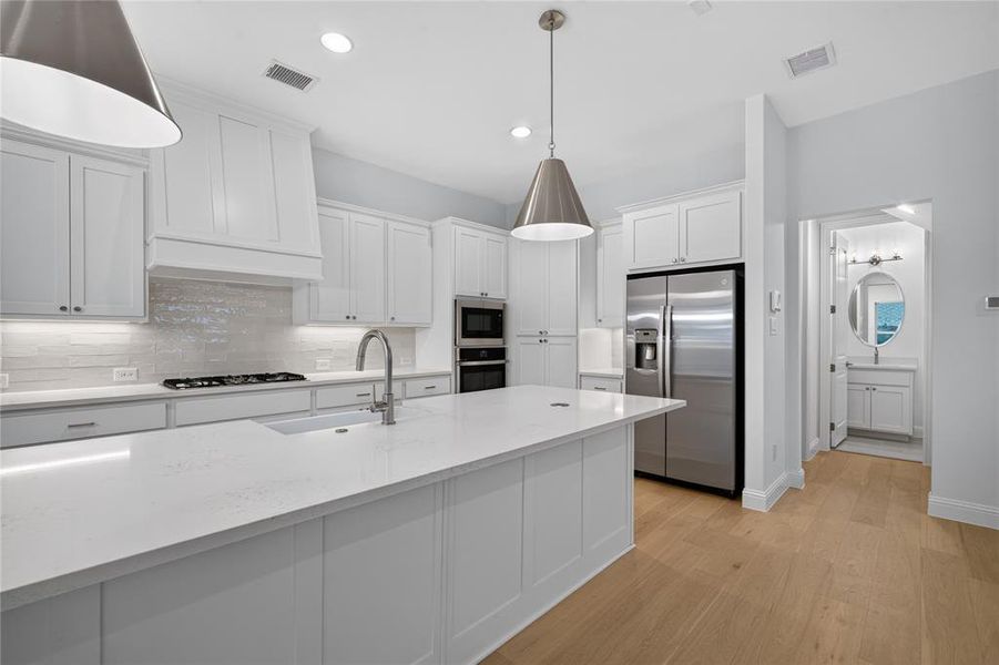 Kitchen featuring a large central island with an undermount sink, white shaker cabinetry, stainless steel appliances, a tile backsplash, and light wood-finish flooring Kitchen featuring a large central island with an undermount sink, white shaker cabinetry, stainless steel appliances, a tile backsplash, and light wood-finish flooring