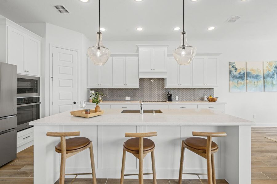 A spacious island with seating for three, pendant lighting, and sleek stainless steel appliances. Bright white cabinetry and a stylish herringbone backsplash enhance the contemporary look, while the wood-look tile flooring adds warmth.