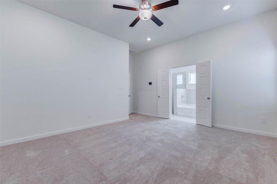 Primary bedroom view showcasing the private en-suite bath entry, plush carpet, high ceiling, ceiling fan, and recessed lighting. The room feels open and serene, with a flexible wall layout for furniture placement and direct access to the bathroom for added everyday convenience.