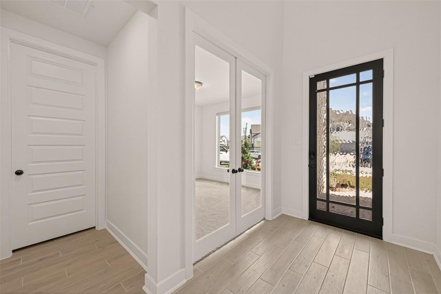 This entryway features a modern glass front door with textured panels, offering a stylish entrance. It leads to a bright room with double glass doors, and has light wood-look tile flooring throughout for a sleek, contemporary look.