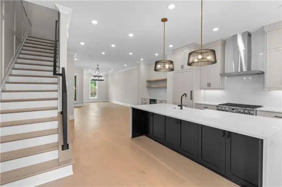 Open-concept layout featuring wide plank wood-finish flooring, a kitchen island with a light-toned countertop, a professional-grade range with a stainless steel vent hood, and a staircase with a black metal railing