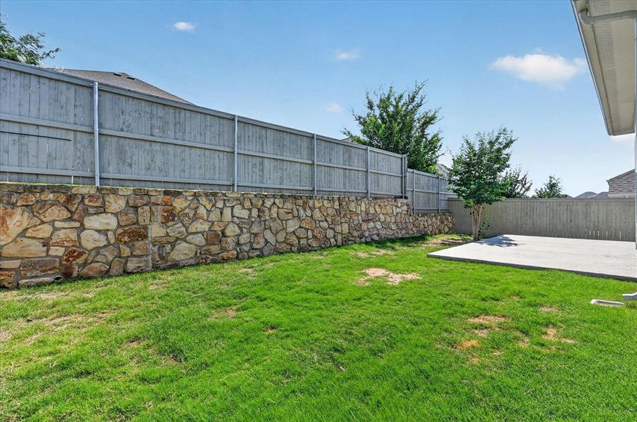 Expansive backyard featuring a stone retaining wall, wood privacy fencing, a concrete patio, and a grass lawn Expansive backyard featuring a stone retaining wall, wood privacy fencing, a concrete patio, and a grass lawn