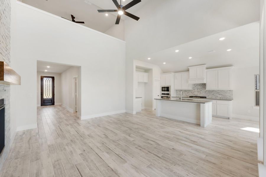 Open-concept living area with high ceilings, ceiling fan, light wood floors, and kitchen with white cabinets and island. Open-concept living area with high ceilings, ceiling fan, light wood floors, and kitchen with white cabinets and island.
