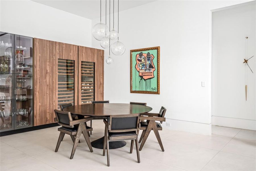 This stylish dining area is framed by custom Madeval cabinetry and floating shelves, featuring glass-fronted display cabinets and dual Subzero wine fridges for easy entertaining. The space is beautifully  illuminated by modern globe pendants and sleek recessed lighting.