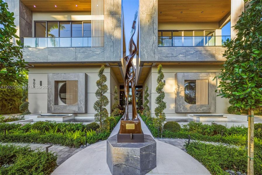 Close-up of paver entry walkway centered on large-scale bronze figurative sculpture on stone pedestal, flanked by spiral topiaries, reflecting pools, stone and stucco facade, wood-ceiling portico, and glass balconies above.