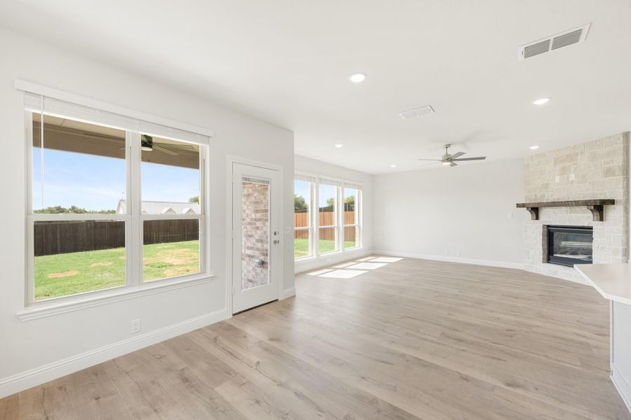 Open living room with light wood floors, stone fireplace, ceiling fan, and large windows overlooking backyard