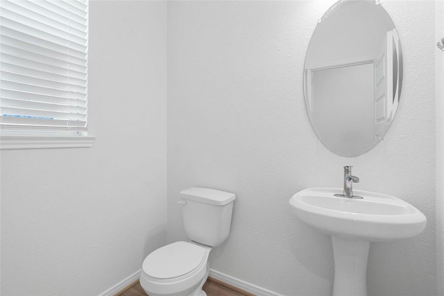 The powder room on the first floor includes a window with privacy blinds, pedestal sink, mirror, and toilet to conveniently meet your needs.