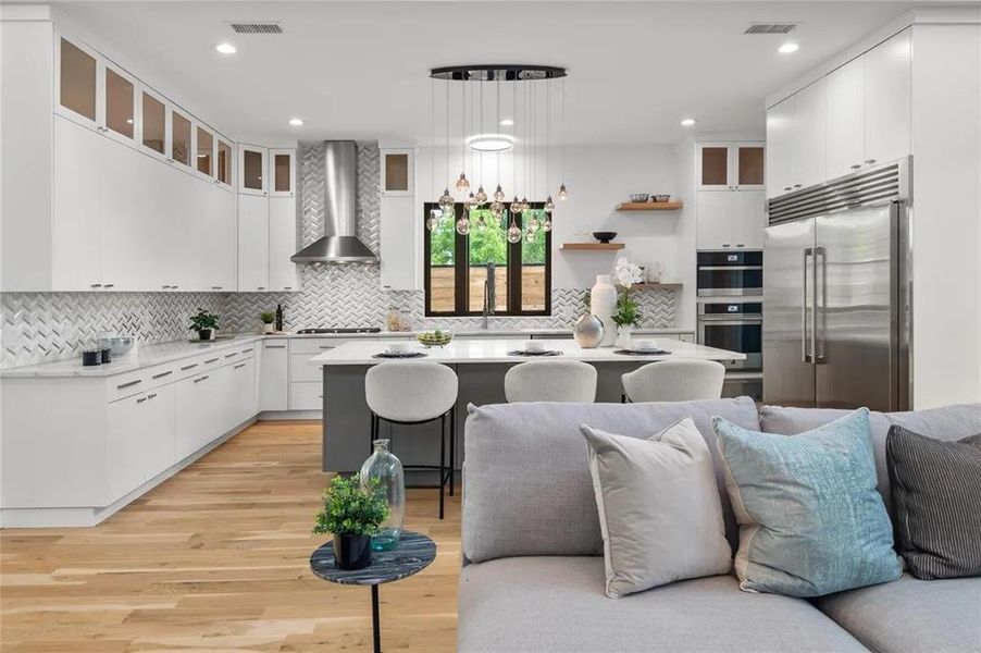 Open-concept kitchen featuring an oversized island with seating, a chevron-patterned tile backsplash, stainless steel appliances, abundant white cabinetry with glass upper accents, and wood-finish flooring Open-concept kitchen featuring an oversized island with seating, a chevron-patterned tile backsplash, stainless steel appliances, abundant white cabinetry with glass upper accents, and wood-finish flooring